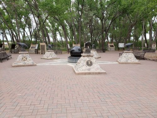 A plaza with stone monuments and sculptures surrounded by trees, featuring benches and informational displays.
