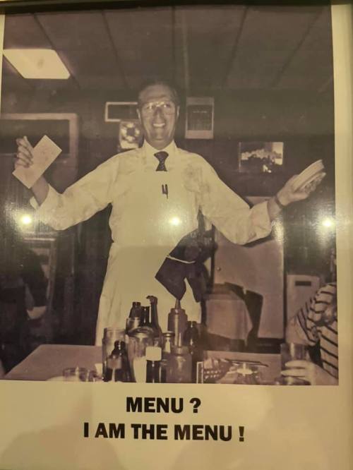 A smiling waiter in an apron holds a menu, playfully declaring, "Menu? I am the menu!" in a vintage restaurant setting.
