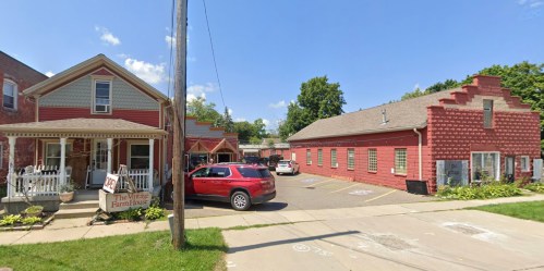 A red building and a house with a porch, surrounded by green grass and a clear blue sky. Cars are parked nearby.