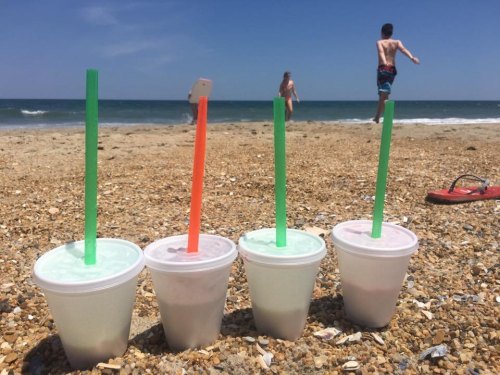 Four cups with colorful straws sit on a sandy beach, with children playing in the background and the ocean in view.