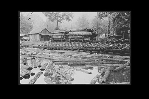 Black and white image of logs stacked near a river, with a wooden structure in the background and trees surrounding the area.