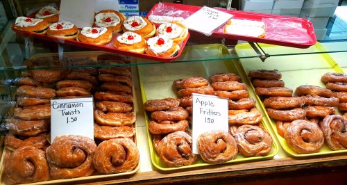 A display case filled with cinnamon twists and apple fritters, labeled with prices, in a bakery setting.