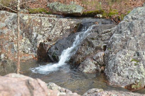 A small waterfall cascades over rocky terrain, surrounded by trees and moss in a serene natural setting.