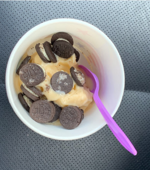A cup of ice cream topped with crushed Oreo cookies and a purple spoon, resting on a textured surface.