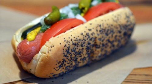 A poppy seed hot dog bun filled with a sausage, topped with tomatoes, onions, pickles, and mustard.