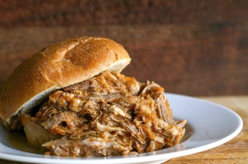 A pulled pork sandwich on a whole grain bun, served on a white plate against a wooden background.
