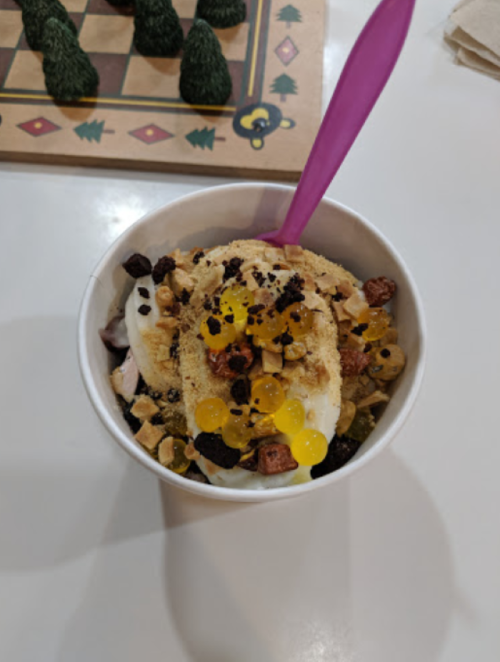A cup of frozen yogurt topped with crushed cookies, candy pieces, and colorful fruit pearls, with a purple spoon.