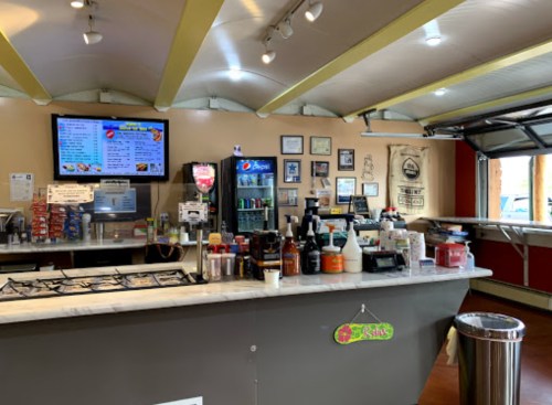 A cozy café counter with a menu screen, drinks fridge, and various condiments displayed on the countertop.