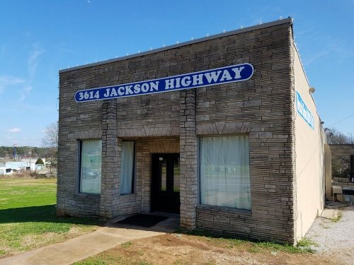 Exterior of a stone building with a sign reading "3614 Jackson Highway" and large windows on a grassy lot.