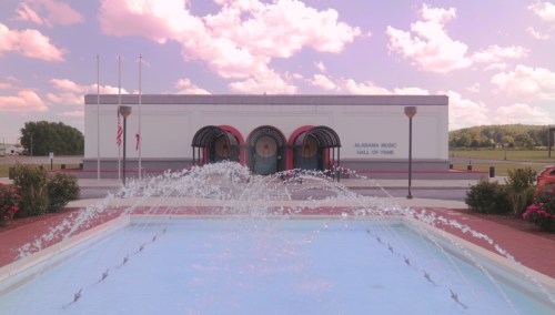 Exterior view of the Alabama Music Hall of Fame, featuring a fountain and colorful clouds in the background.