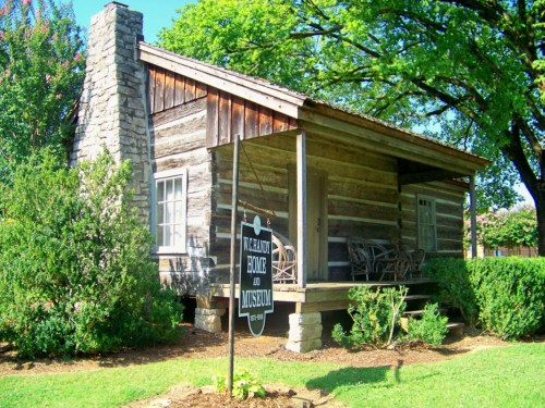 A rustic log cabin with a stone chimney, surrounded by greenery, featuring a sign for the home and museum.