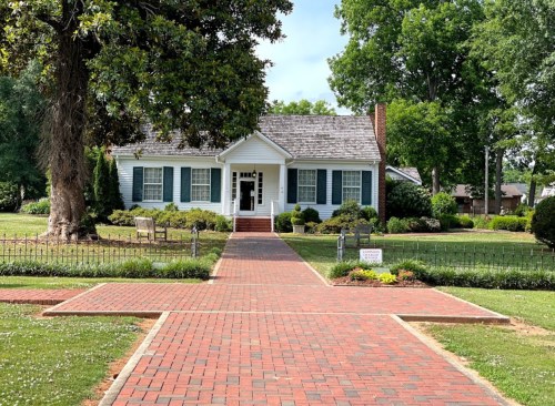 A charming white house with green shutters, surrounded by lush greenery and a brick pathway leading to the entrance.