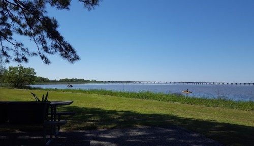 A serene lakeside view with a bridge in the distance, kayakers on the water, and green grass in the foreground.