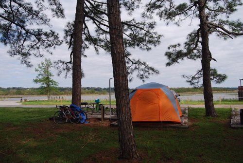 A camping scene featuring an orange tent nestled between trees, with bicycles nearby and a view of a lake in the background.