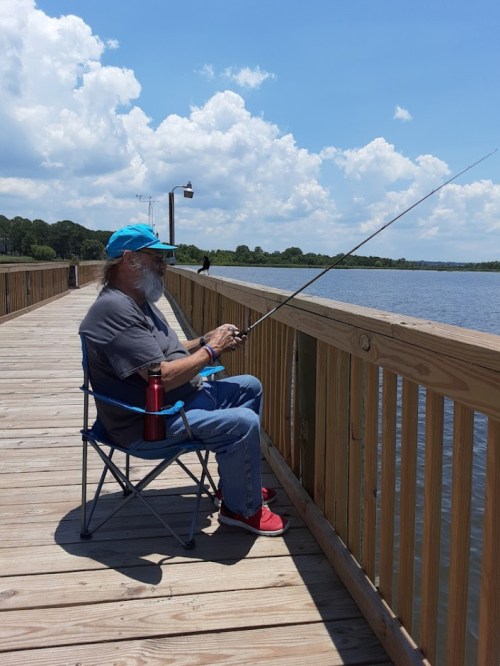An older man sits on a dock, fishing with a rod, wearing a blue cap and enjoying a sunny day by the water.
