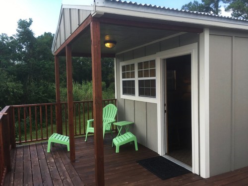 A small porch with green chairs and a table, leading to a door, surrounded by trees and greenery.