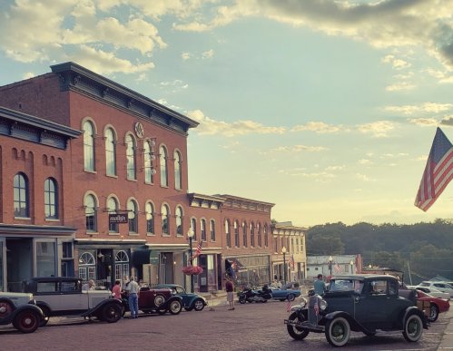 Historic street scene with vintage cars, brick buildings, and an American flag under a sunset sky.
