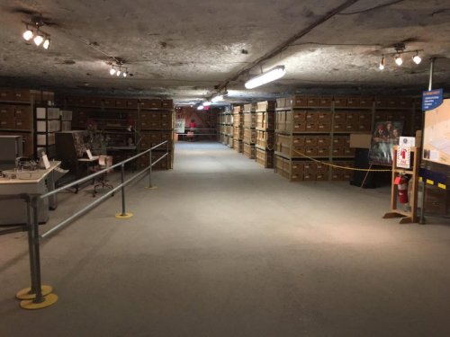 A dimly lit underground storage area with rows of stacked boxes and a railing along the path.