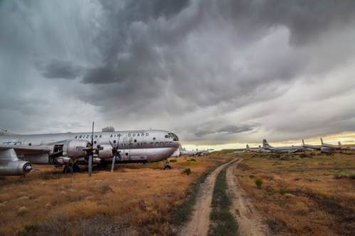 Abandoned military aircraft in a field under a dramatic sky, with a dirt path leading through the overgrown grass.