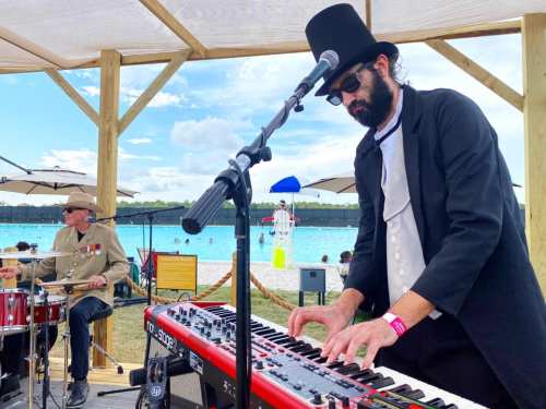 A musician in a top hat plays a keyboard at a beachside event, while a drummer performs nearby.