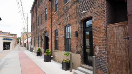 A brick building with large windows and planters, alongside a paved walkway and alleyway.