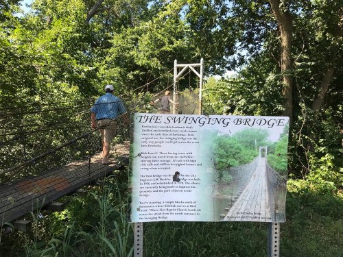 A person walks across a swinging bridge surrounded by greenery, with an informational sign nearby.