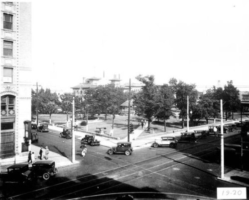 Historic black-and-white photo of a busy street corner with vintage cars and trees in a park, dated 1920.