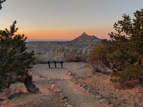 A scenic view of a path leading to a bench, with rocky formations and a distant castle-like structure at sunset.