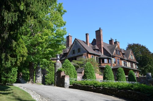 A large, historic mansion with a stone driveway, surrounded by lush greenery and tall trees under a clear blue sky.