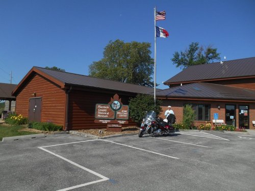 A wooden building with a sign for the Harrison County Historical Museum, featuring flags and a motorcycle parked outside.
