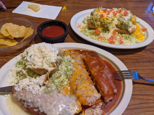 A plate of enchiladas with cheese and sauce, served with lettuce and a side of chips and salsa. Another plate with tacos.