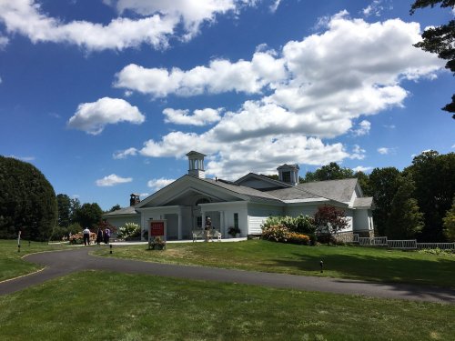 A white building with a sloped roof and two cupolas, surrounded by green grass and colorful flowers under a blue sky.