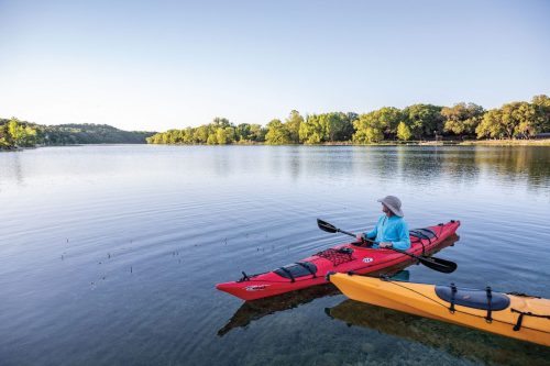 A person in a red kayak sits on calm water, surrounded by trees and a clear sky, with a yellow kayak nearby.