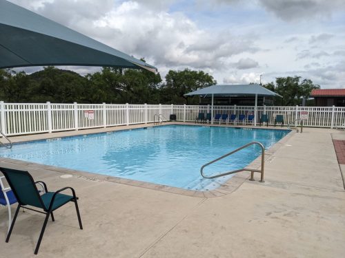 A clear swimming pool surrounded by a white fence, with shaded seating areas and cloudy skies above.