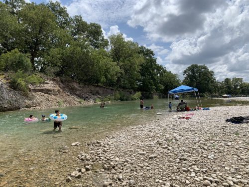 People enjoying a sunny day by a river, swimming and relaxing under a blue tent, surrounded by trees and rocky shore.