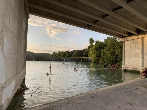 A serene scene under a bridge, with people swimming and playing in the water as the sun sets in the background.