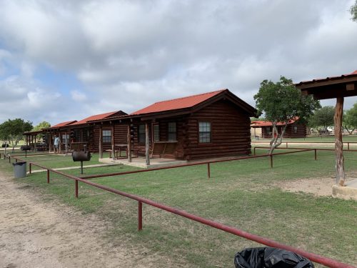 Log cabins with red roofs in a grassy area, surrounded by trees and a cloudy sky.