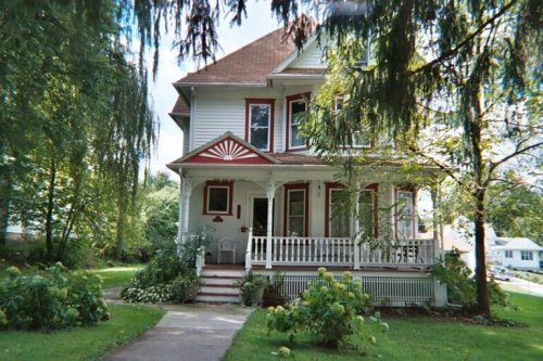 A charming two-story house with a porch, surrounded by greenery and trees on a sunny day.