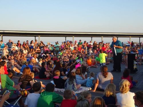 A large crowd gathers on a boardwalk for a performance, with a magician entertaining children and families.