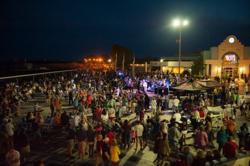 A large crowd gathers at night for a concert on a boardwalk, with lights and a stage in the background.