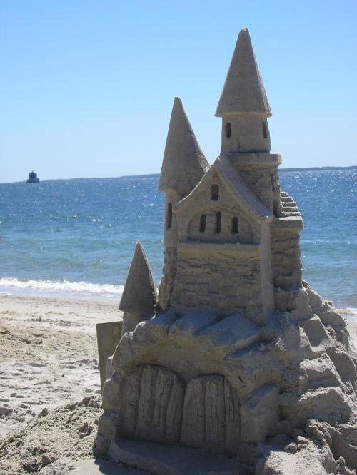 A detailed sandcastle with towers sits on a beach, with the ocean and a distant ship in the background.