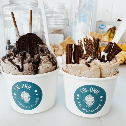 Two cups of rolled ice cream topped with chocolate, cookies, and sticks, with a logo reading "Tai-Okie."
