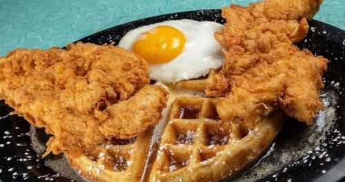 A plate featuring crispy fried chicken, a sunny-side-up egg, and a waffle drizzled with syrup.