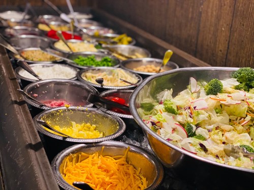 A variety of salad toppings in metal bowls, including vegetables, cheese, and dressings, arranged on a buffet table.