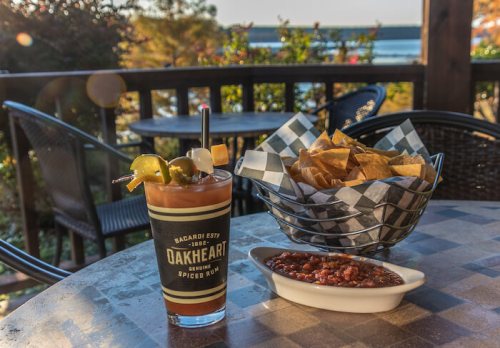 A cocktail garnished with pickles and a skewer, served with chips and chili, with a scenic view in the background.