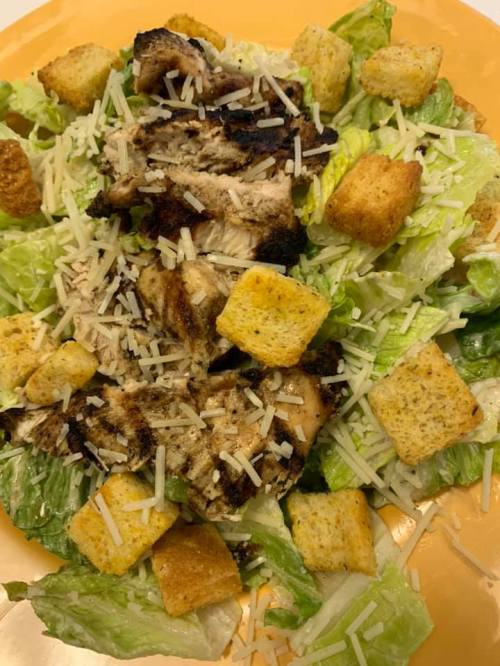 A plate of Caesar salad topped with grilled chicken, croutons, and shredded cheese on a bed of romaine lettuce.