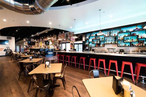 A modern bar interior featuring a long counter, colorful shelves, and tables with chairs in a vibrant setting.
