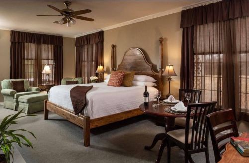 Cozy hotel room with a king bed, seating area, and warm lighting from lamps and windows. Elegant decor and a dining table.