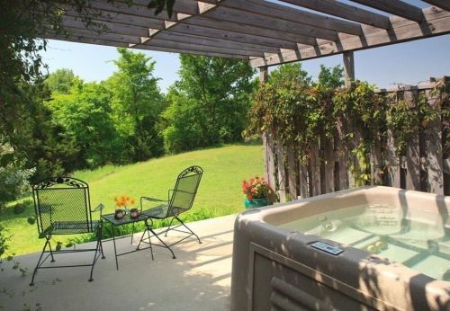 A serene outdoor patio with a hot tub, two chairs, and lush greenery in the background under a wooden pergola.