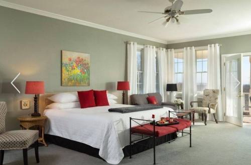 A cozy bedroom with a large bed, decorative pillows, a small table, and bright windows overlooking a scenic view.